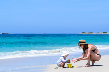 Young cute mother in sunglasses, straw hat and swimwear and her adorable little daughter in swimsuit building sandcastles at tropical white sandy beach during vacation over background of turquoise sea