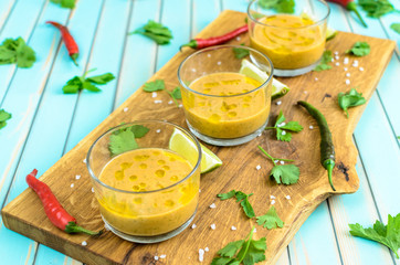 Cold fresh homemade tomato soup gazpacho in glasses on cutting board and ingredients with red, yellow tomatoes, celery, lime and chili peppers over rustic turquoise wooden background, close-up