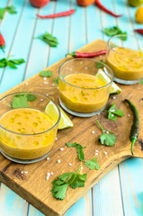 Cold fresh homemade tomato soup gazpacho in glasses on cutting board and ingredients with red, yellow tomatoes, celery, lime and chili peppers over rustic turquoise wooden background, close-up