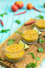 Cold fresh homemade tomato soup gazpacho in glasses on cutting board and ingredients with red, yellow tomatoes, celery, lime and chili peppers over rustic turquoise wooden background, close-up