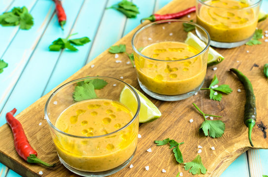 Cold fresh homemade tomato soup gazpacho in glasses on cutting board and ingredients with red, yellow tomatoes, celery, lime and chili peppers over rustic turquoise wooden background, close-up
