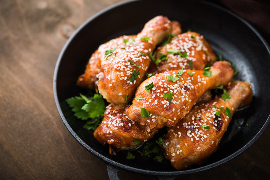 Baked spicy chicken legs with sesame and parsley in cast iron frying pan on dark wooden background close up.