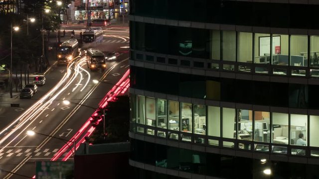Timelapse shot of night car traffic with modern office business centre in foreground, people working in the office can be seen through the windows. Seoul, South Korea