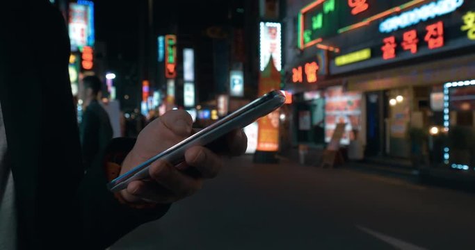 Close-up shot of woman scrolling up and down on smart phone. Night street of Seoul with illuminated banners in background, South Korea