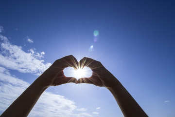 Heart shaped by the hands of a beautiful woman on bright sky background and sunlight with flare in the afternoon.