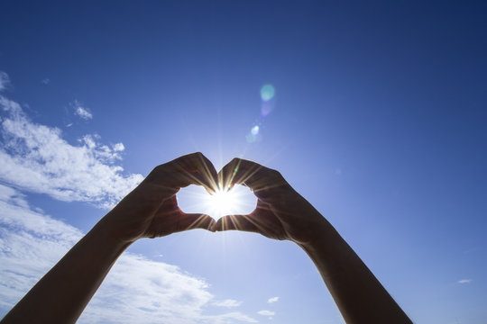 Heart shaped by the hands of a beautiful woman on bright sky background and sunlight with flare in the afternoon.