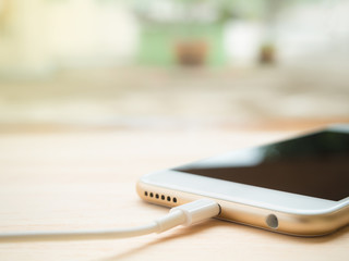 Close-up of white smartphone charging battery with cable on wooden table with copy space and blurry background. Soft light