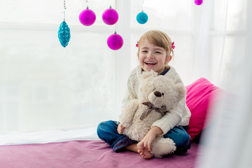 Pretty little girl sitting on windowsill and embraces a teddy bear. New Year's window decorated with colored balls