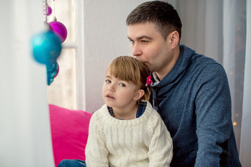 Dad kissing little daughter sitting on the windowsill. Window decorated with colored balls New Year