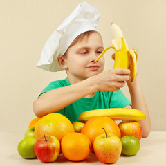 Little boy in chefs hat peeling fresh banana at the table with fruits