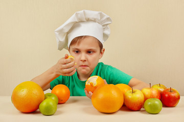 Little funny chef peeling fresh orange at the table with fruits