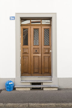 Frontansicht einer eleganten, braunen Haustür mit blauem Hausnummernschild auf der linken Wandseite und einem blauen Abfallsack auf dem Boden links von der Tür.