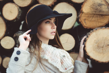 Gorgeous young woman in coat and hat posing by tree chunks in autumn