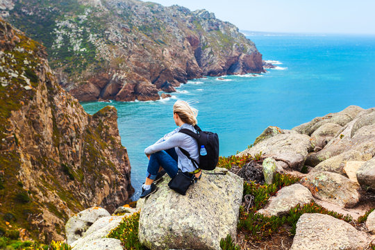Female traveler sitting on Cape Roca against the background of the Atlantic Ocean, Sintra, Portugal