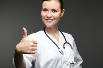 Portrait friendly, smiling beautiful doctor with stethoscope on grey background. She shows thumb up sign. Selective focus. Positive face expression emotion attitude.