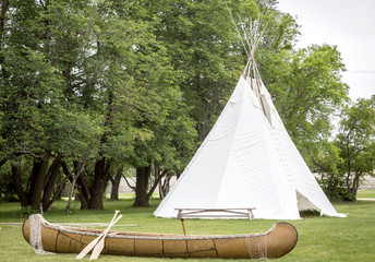horizontal image of a white teepee sitting on green grass surrounded by green trees with a brown...