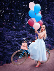 Outdoor portrait of a beautiful blonde girl holding balloons and riding a bike