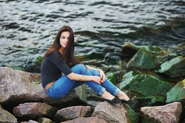 Beautiful young dark-haired girl in jeans and a black t-shirt posing on the rocks in the sea, photo shoot on the beach.
