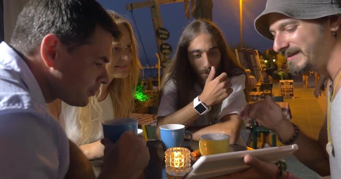 In evening in city of Perea, Greece at the table in cafe sits a young company of 3 men and one girl. One of the men on tablet shows to other people photo