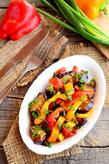 Stewed vegetables on a plate and on old wooden background. Peppers with aubergine, garlic and green onions. Ingredients for cooking vegetable stews, fork, knife. Hearty vegetable food. Top view