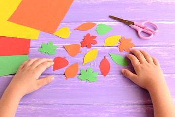 Child cut out of colored paper autumn leaves. Kid doing autumn paper crafts. Children hands, scissors on lilac wooden background. Hand made autumn diy for kids. Preschool education art and craft