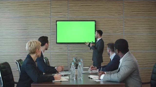 Businessman Standing With Mic In Front Of His Colleagues In The Meeting Room And Explaining Something On Green Screen Of TV