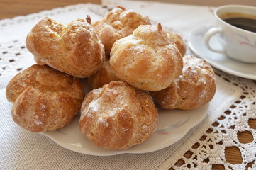 Delicious, small homemade cream puffs powdered with icing sugar on white plate and cup of coffee
