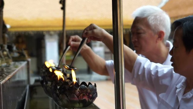 Asian senior doing Buddhist ritual pouring oil to fill candle frame for BUddha statue