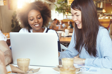 Young female friends having coffee break, sitting at cafe in front of laptop with copy space for your text or promotional content, using computer for sharing ideas on common business project