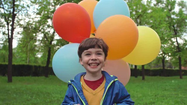 Dolly shot of little preschooler with wide toothy smile walking through green park holding bunch of colorful balloons and looking at camera
