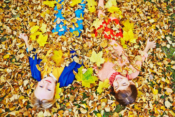 Above view of two happy children, boy and girl, in bright clothes enjoying warm autumn day, playing in park and lying in pile of fallen leaves throwing them on themselves, blond boy looking at camera