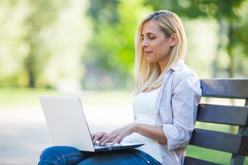 Young happy woman is sitting in park and using laptop.