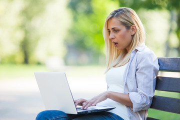 Young woman is sitting in park and reading news on laptop. She is surprised.