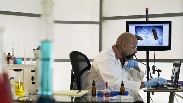 A bearded scientist working in his laboratory moves from electronic computerized microscope to a standard one.