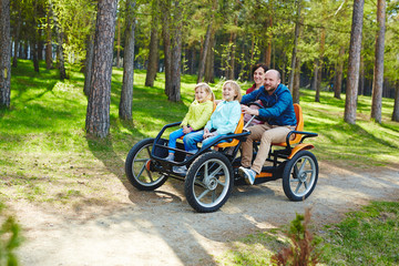 Active family with kids, mother father and two daughters, enjoying ride in four wheeled bicycle car around green forest during weekend entertainment in park