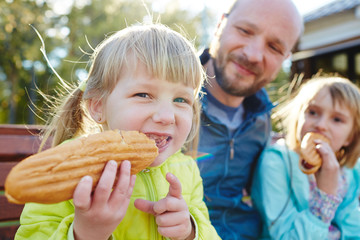 Two blond girls sitting in sunlight on bench with father and enjoying big spicy hot dogs on warm autumn day in park