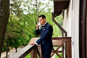 Elegant groom is drinking coffee on the balcony