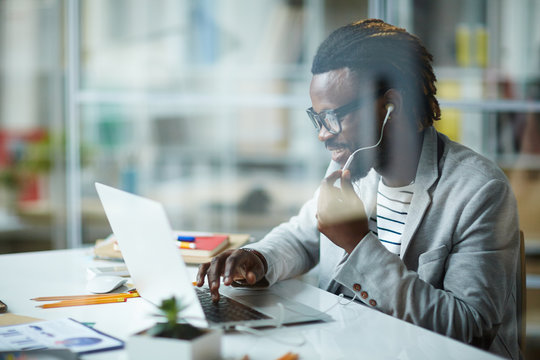 Modern Young African-American Entrepreneur Working At Successful Start-up Business In Small Office, Talking With Partners Using Phone Headset And Laptop, Behind Glass Wall Shot