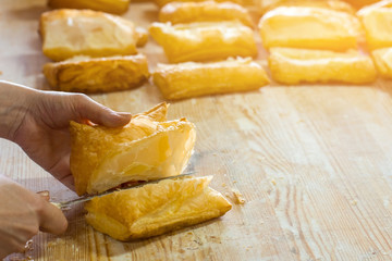 Hand with knife cuts pastry. Yellow puff on wooden board. Crispy and airy dough. Product from the bakery.