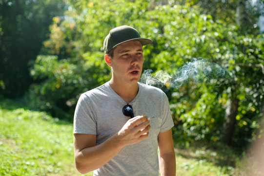 a young man dressed in light and dark ftbolku cap, smoking a cigarette emitting smoke in the background of summer green sunny day