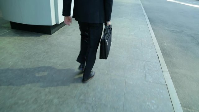 Close crop of businessman feet walking in city. Man is commuting to work, he holds a business briefcase in his hand (steadicam shot)
