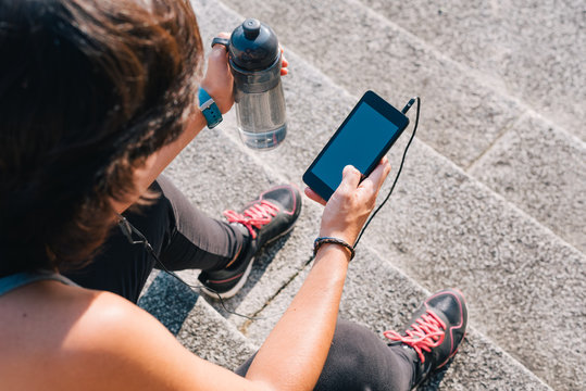 Athlete woman sitting on stone stairs resting and listening music on smart phone with bottle of water while resting from outdoors urban training exercise workout. Female wellness concept.