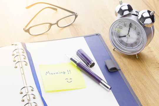 A cup of coffee and opened notebook with MORNING message on wooden background with pen, glasses and flowers. warm tone.