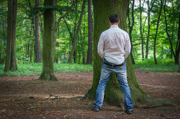 Standing man peeing near big tree in summer forest