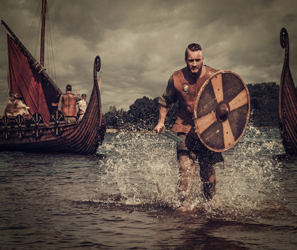 Viking warrior in the attack, running along the shore with Drakkar on the background.