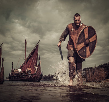 Viking warrior in the attack, running along the shore with Drakkar on the background.
