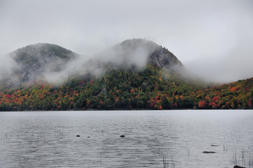 Lake in the mountains, colors around autumn trees.   Acadia National Park. Maine. USA. Cloudy gloomy weather, fog and clouds over the mountains in the distance.