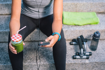 female holding juice vegetable green detox cleanse smoothie after fitness running workout listening music using her phone with bottle of water and towel. Healthy lifestyle concept.