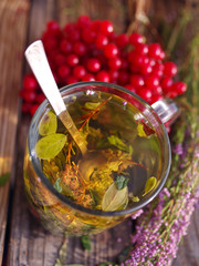 Herbal tea in a transparent glass mug, berries of a guelder-rose and forest herbs on a wooden surface of a table. Tea from autumn herbs and berries. Phytotea   