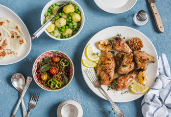 Lunch table. Lemon thyme baked chicken, boiled potatoes with green peas, salad with lentils and tomatoes, flatbread on a blue background. Top view